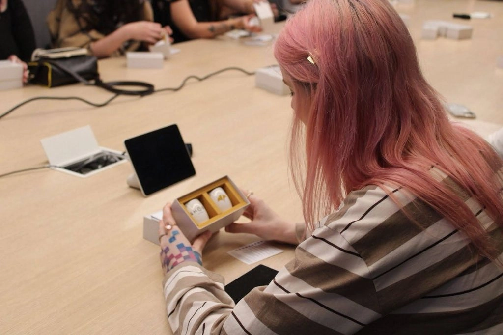 Person with pink hair examines a small box at a meeting table, surrounded by tech devices and people
