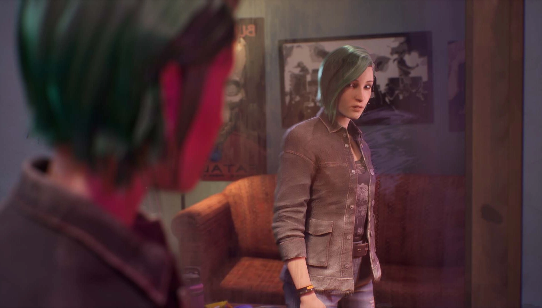 Chloe with green hair looks at her reflection in a mirror, standing in a room with a brown couch