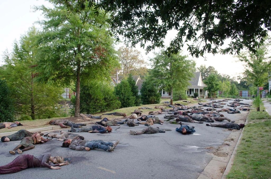 A street littered with motionless zombies in a suburban neighborhood, trees lining the road
