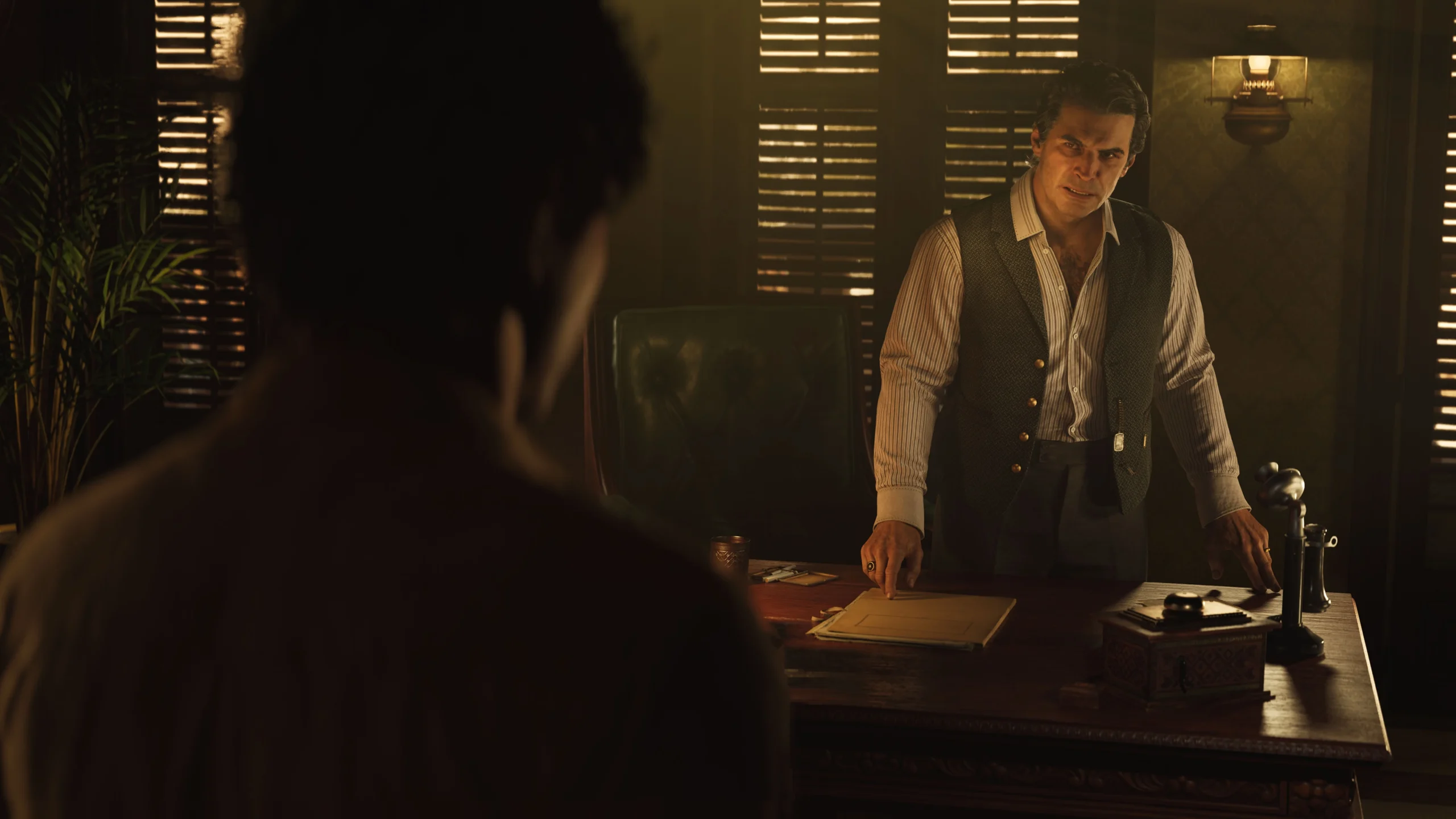 A man in a dimly lit office stands behind a desk, engaged in a serious conversation with another person
