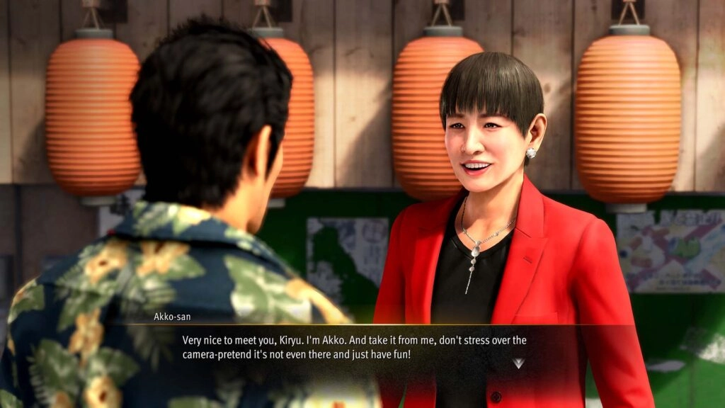 Kiryu talking to Akko-san in a vibrant setting with lanterns, discussing camera presence and having fun