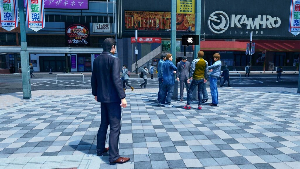 A man in a suit observes a group of people gathered outside Kamuro Theater in a bustling urban area