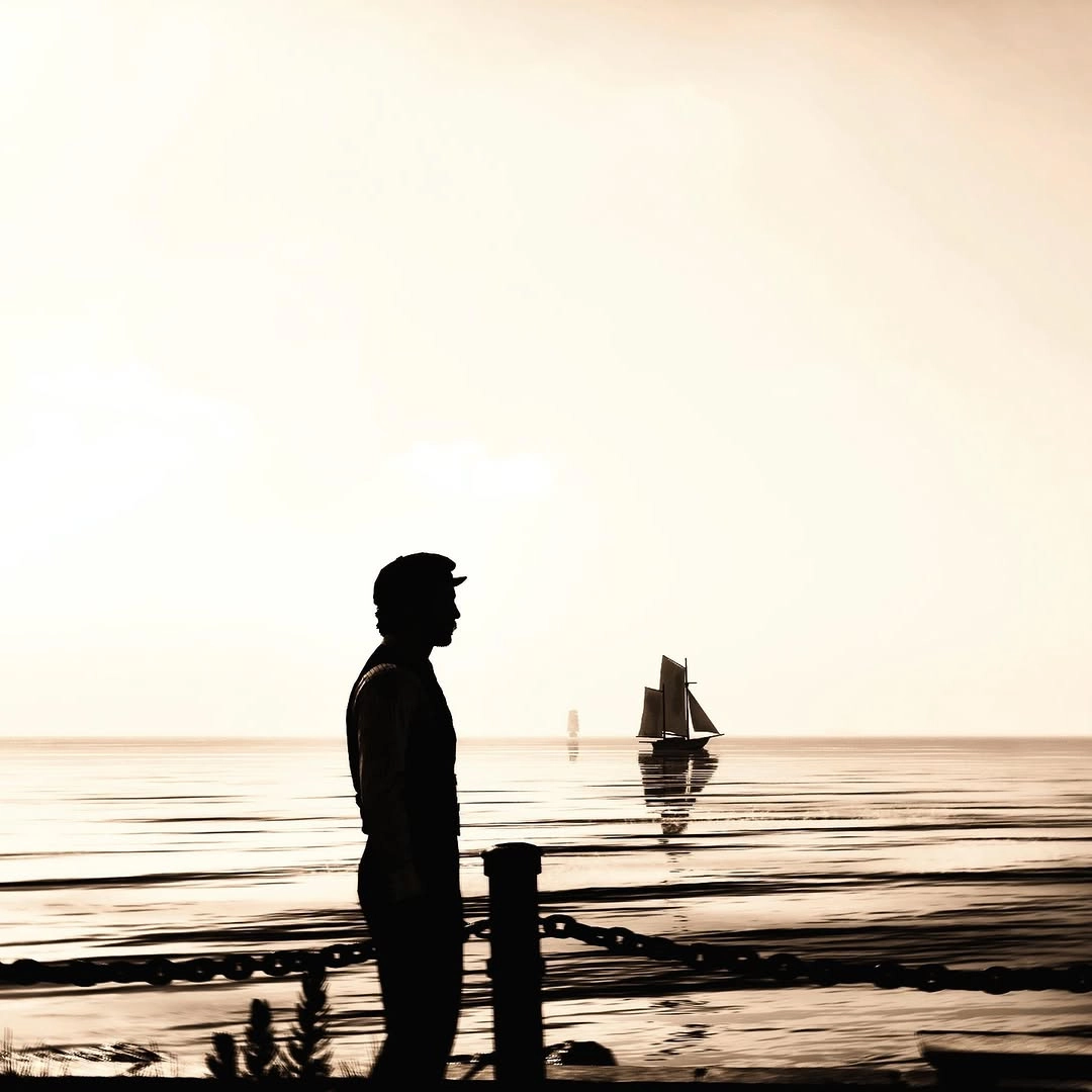 Silhouette of a man by the sea with a distant sailboat, capturing a serene, nostalgic atmosphere