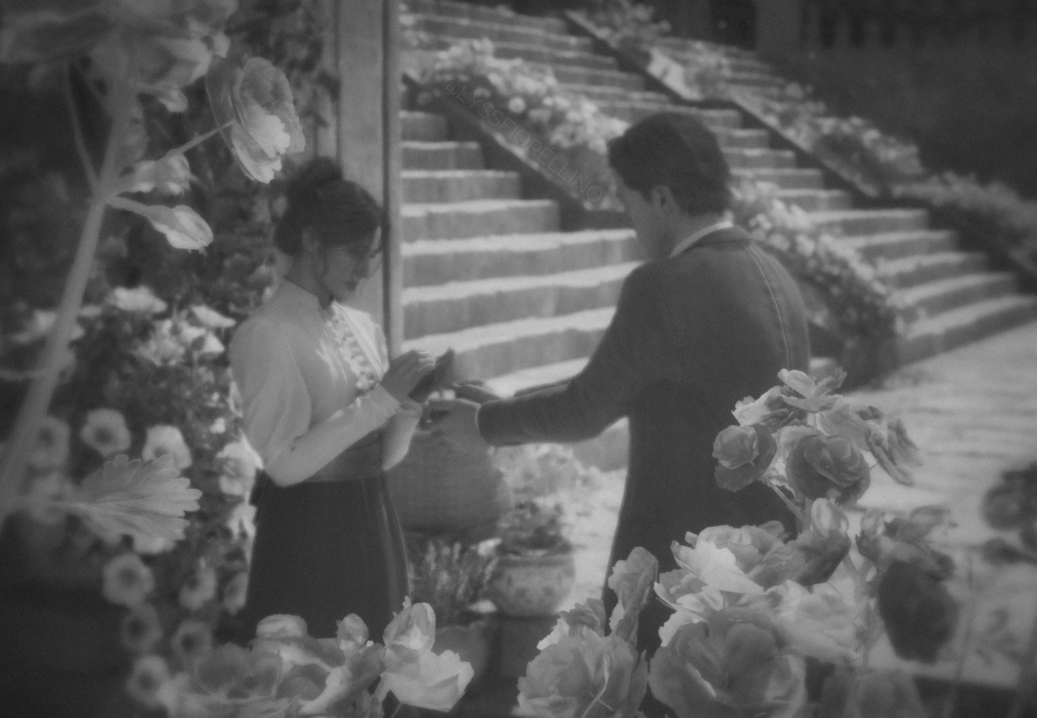 A couple exchanging rings in a garden setting, surrounded by blooming flowers and stone steps