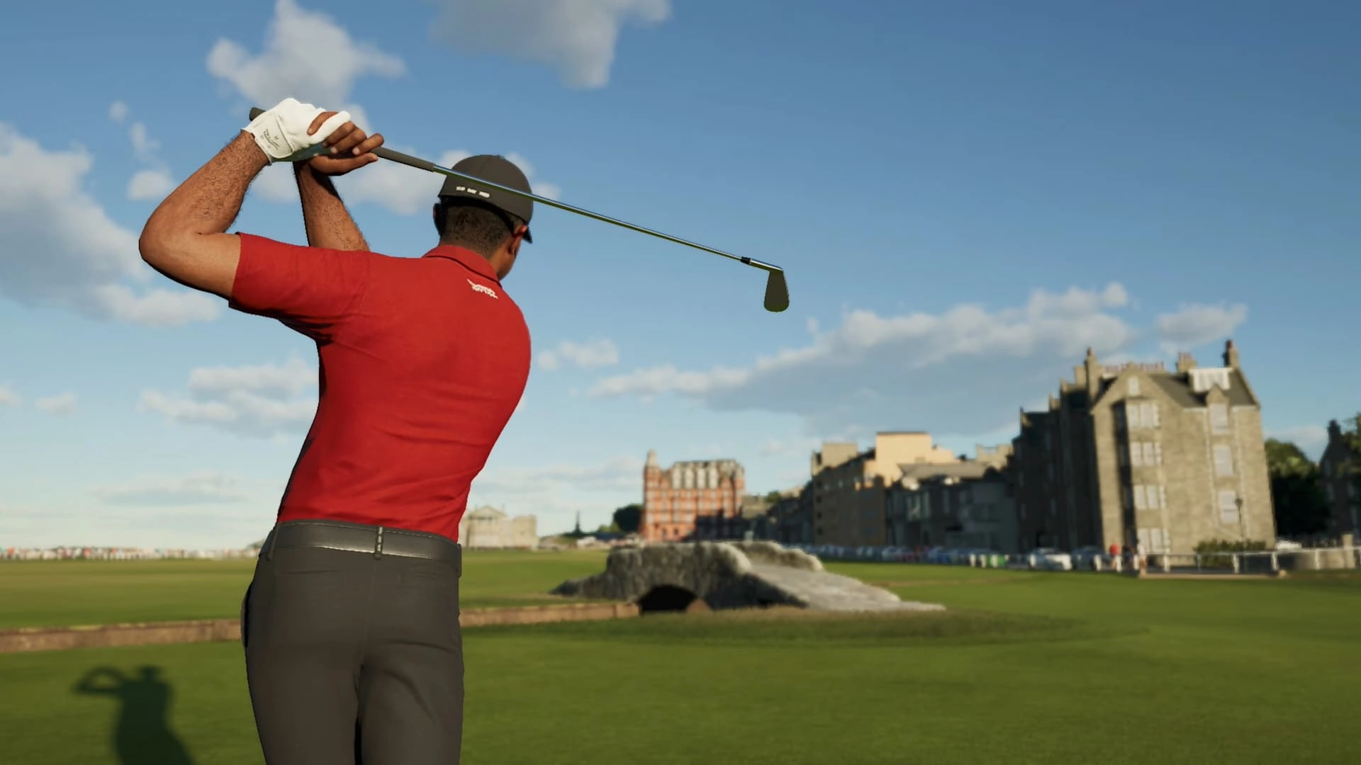 Golfer in red shirt swings club on a lush course, iconic bridge and historic buildings in the background