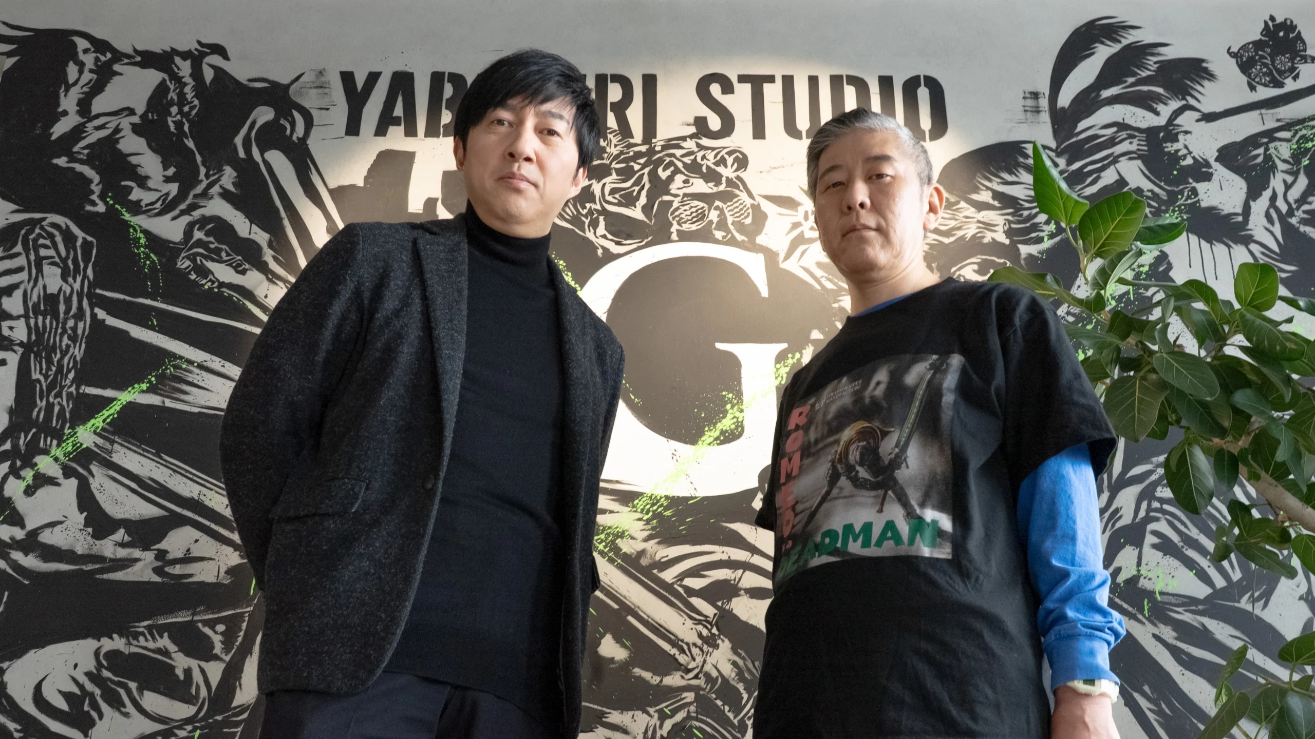 Two men standing in front of a Yabuki Studio sign, one in a black turtleneck, the other in a graphic tee